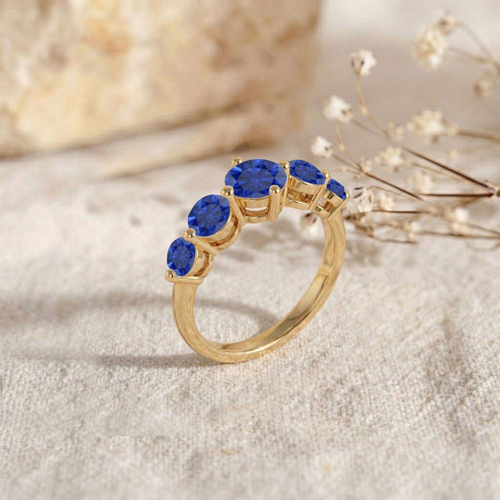 A gold ring featuring five round-cut blue gemstones set in a curved row, displayed on a textured beige fabric with dried baby's breath flowers and a soft-focus stone in the background.