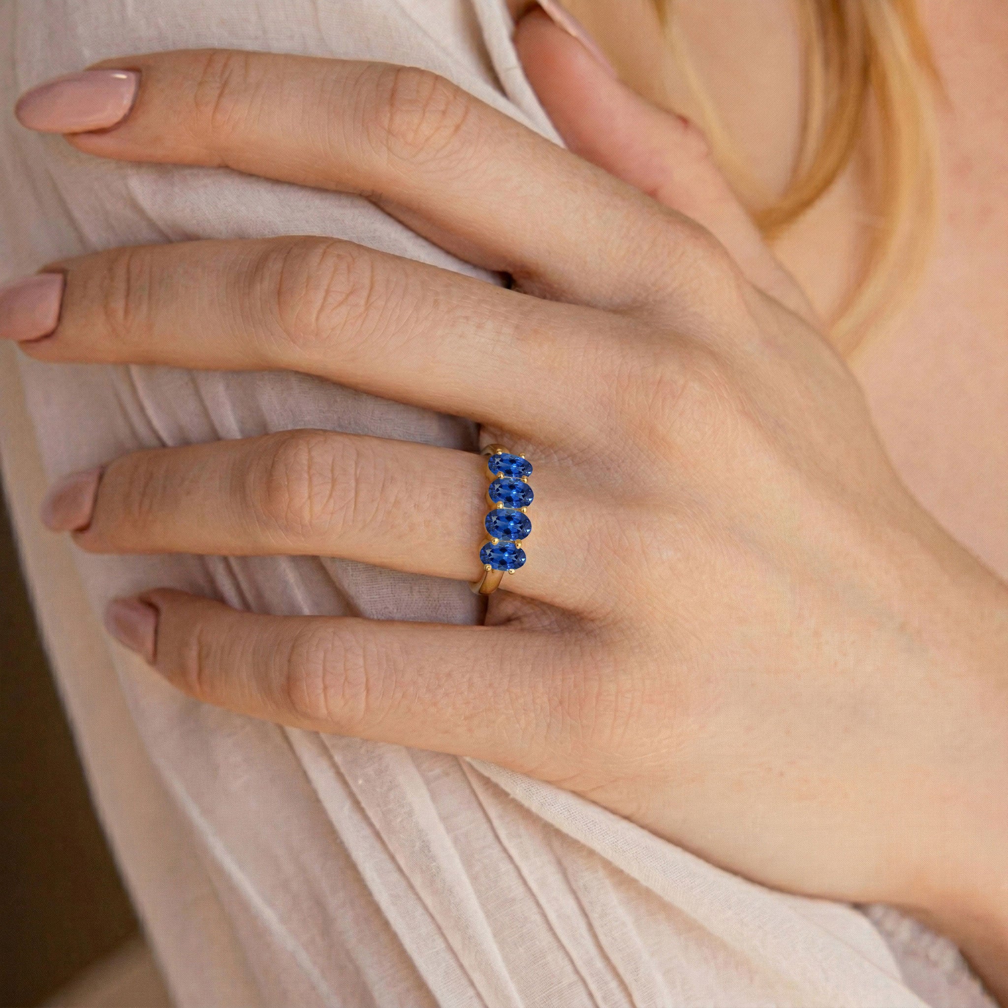 A close-up of a person's hand wearing a gold ring set with five round-cut blue gemstones, resting on a light beige fabric.