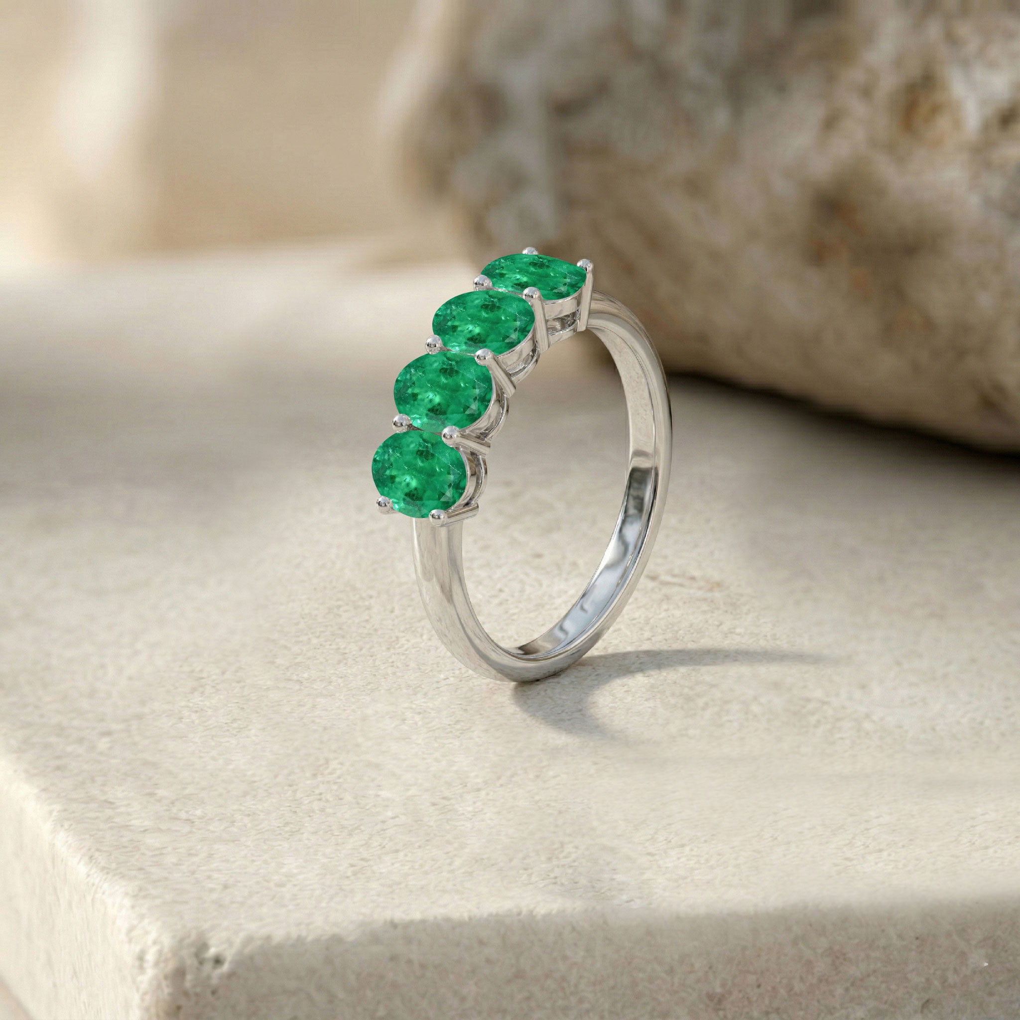 A silver ring featuring four round-cut green emerald-cut gemstones, displayed on a textured beige stone surface.