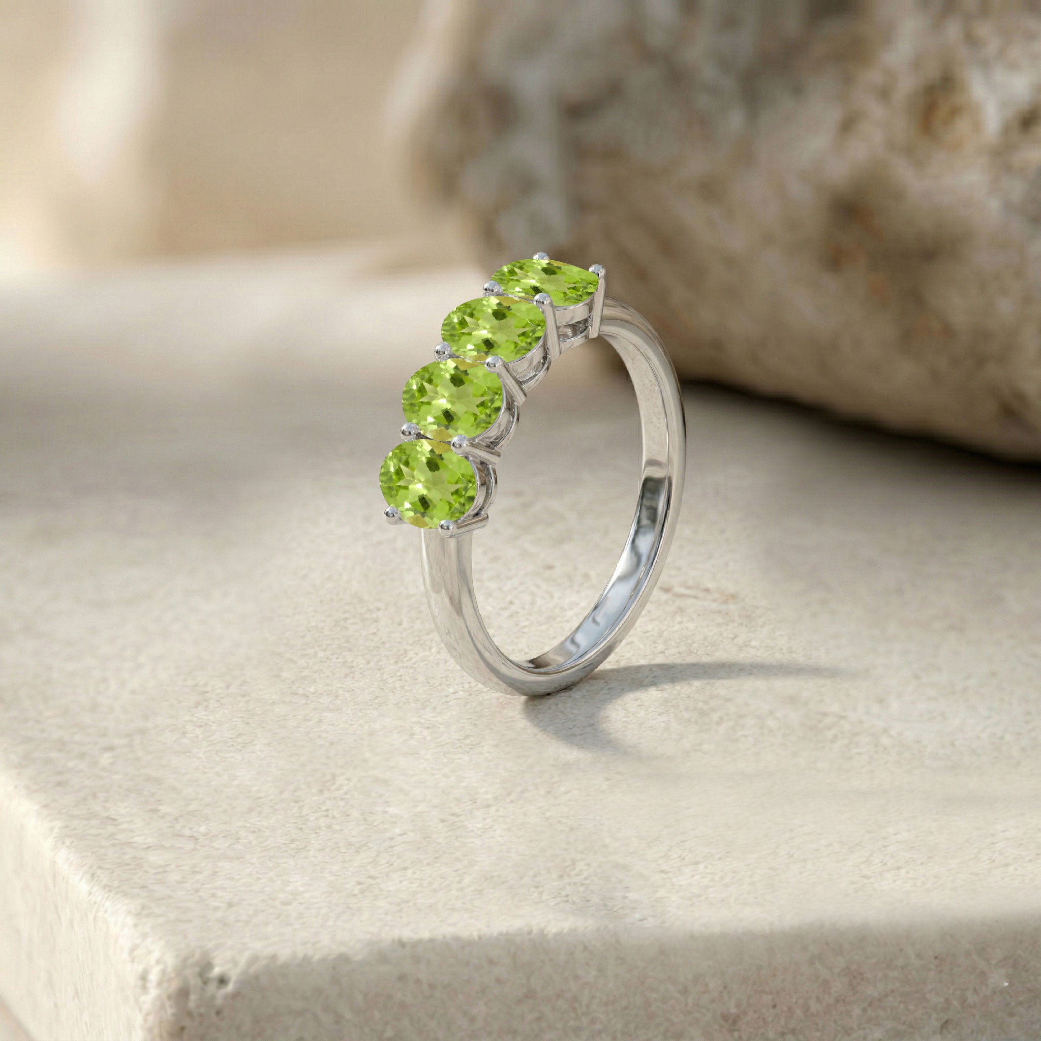 A silver ring featuring four round-cut green peridot gemstones in a row, displayed on a light stone surface with a blurred natural rock background.