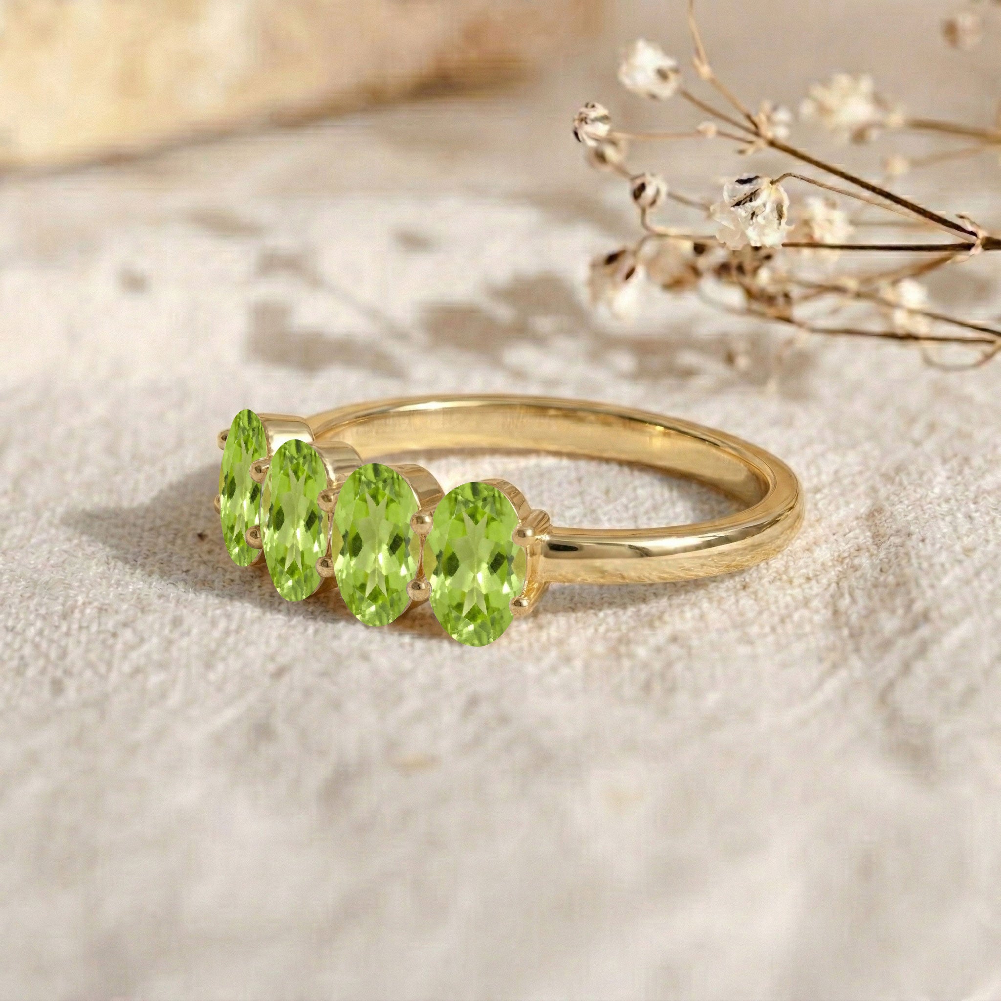 A gold ring featuring four oval-cut light green gemstones, displayed on a textured beige fabric with dried baby's breath flowers in the background.